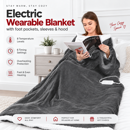 A woman sits in bed reading a magazine while wearing a dark gray electric wearable blanket with foot pockets, sleeves, and a hood. The blanket has a soft plush texture and a white sherpa lining.